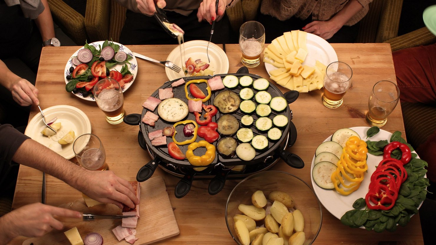 Table with raclette oven and dishes with potatoes, vegetables and cheese